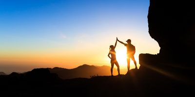 Teamwork couple helping hand trust help, silhouette success in mountains. Team of climbers man and woman. Hikers celebrate with hands up, help each other on top of mountain, climbing together, beautiful sunset landscape.